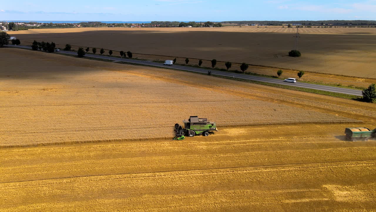 곡물 트레일러가 실린 트랙터가 결합 수확기를 떠나고, 기계는 수확을 계속하고, 들판은 도로에 가깝습니다.
