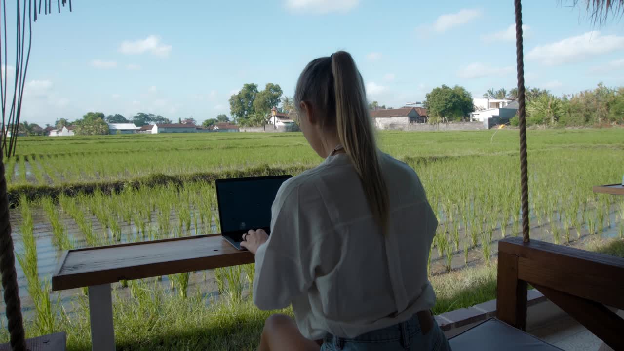 푸른 드레스를 입은 젊은 유럽 소녀가 노트북에서 원격으로 온라인으로 작업하고 초록색 식물과 함께 뒷마당의 화면을보고 있습니다.