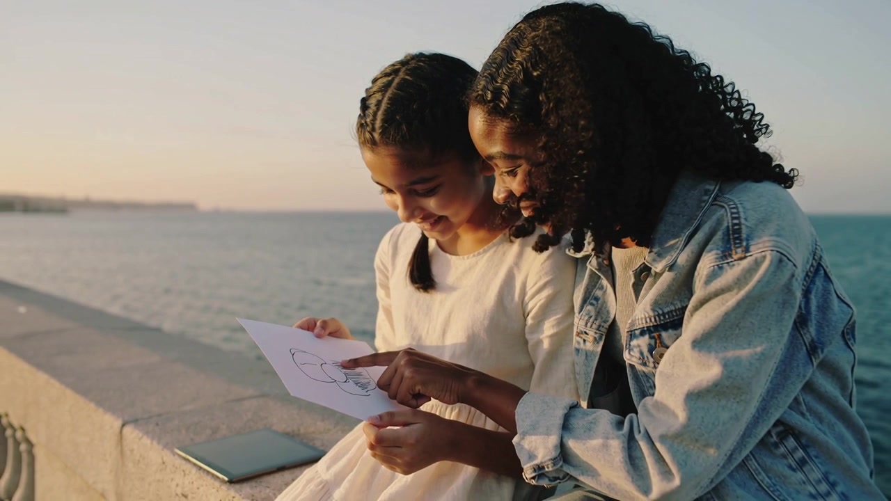 Girl showing drawing, mum leaning and pointing at paper on seawall to praise, denim jacket, tablet