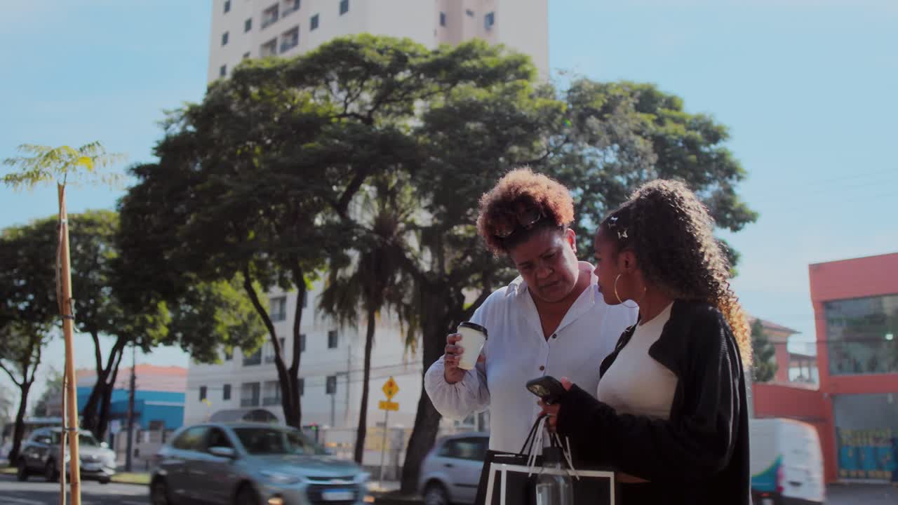 Two women interact on a city street, one using a smartphone and the other holding a coffee cup