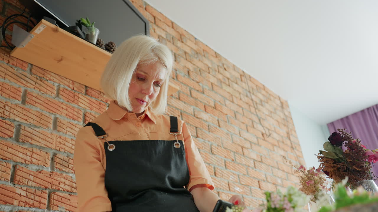 Female florist in black apron photographing floral arrangement with smartphone in rustic workshop, standing against brick wall with dried flowers, purple curtain in background