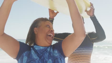 feliz pareja afroamericana de alto nivel caminando con tabla de surf en una playa soleada