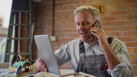 mature male carpenter in garage workshop with digital tablet talking on mobile phone