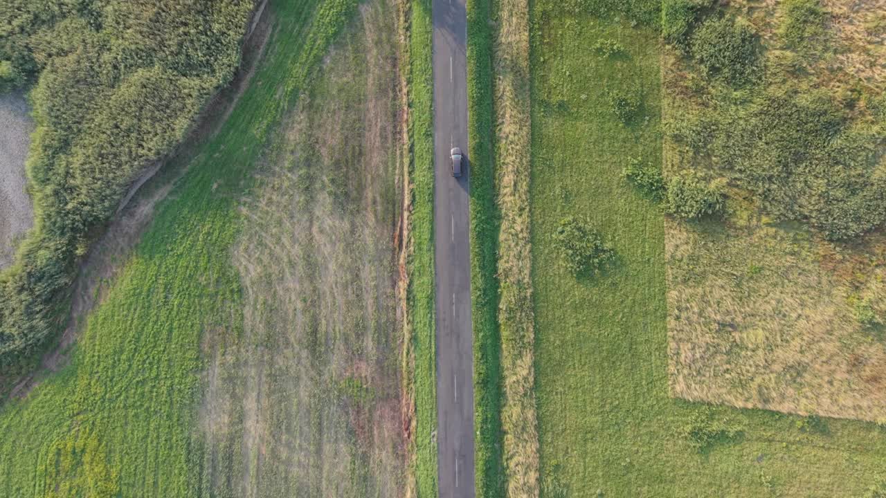A car travels along a perfectly straight rural road surrounded by vast green fields. The aerial perspective highlights the symmetry of the landscape and the peaceful atmosphere of the open countryside