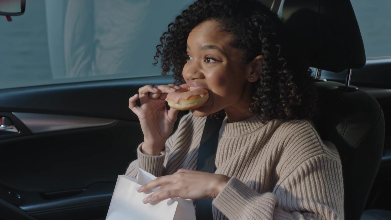 Reaching into white paper bag, woman in tan sweater pulling pink donut, eating at parked car