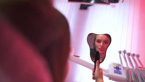 lady vampire enjoys her teeth reflection in the mirror. blonde female dentist nearby. dentistry cabinet lighted in pink backdrop.