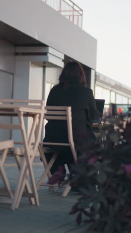 vista trasera de una mujer escribiendo en una computadora portátil, rodeada de sillas de madera y plantas en una cafetería al aire libre, la escena es pacífica con un vistazo de una persona en la distancia caminando