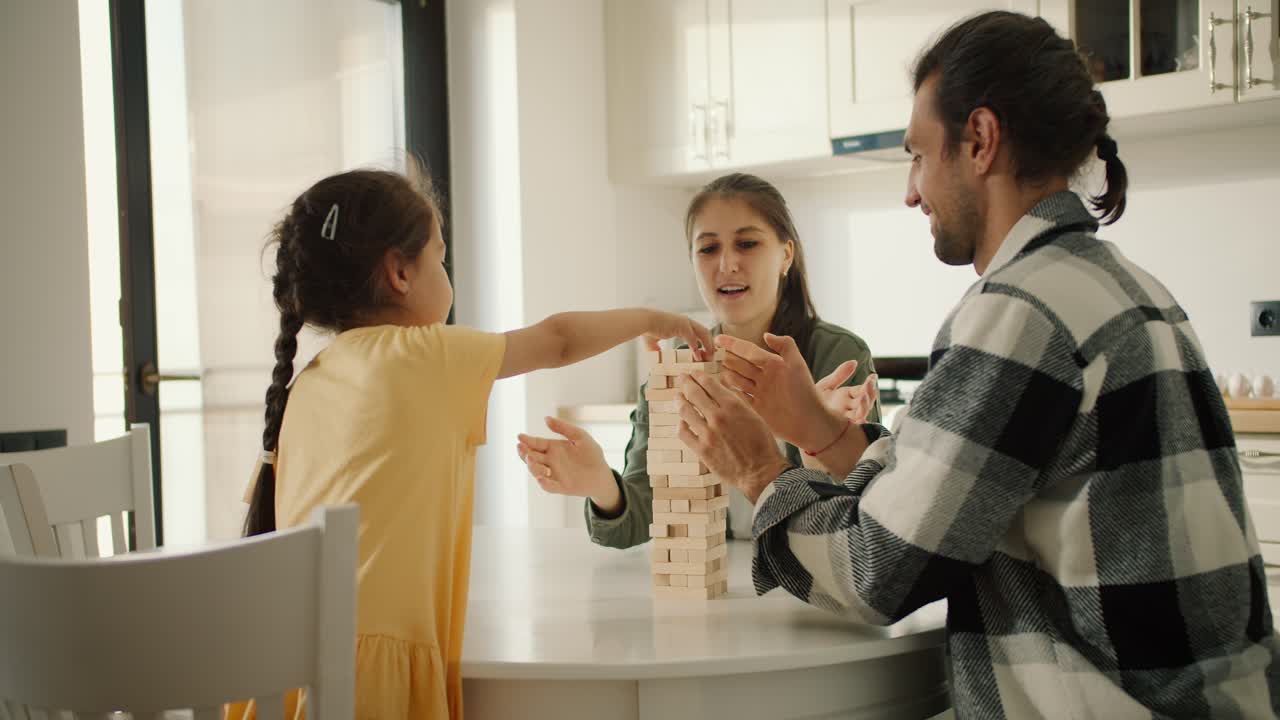 노란색 드레스를 입은 작은 갈색 소녀의 뒷면은 색 현대 부의 테이블에서 부모와 함께 보드 게임 젠가 (jenga) 를 연주합니다.