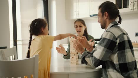 vista trasera de una pequeña chica morena en un vestido amarillo jugando el juego de mesa jenga con sus padres en la mesa en una cocina moderna blanca
