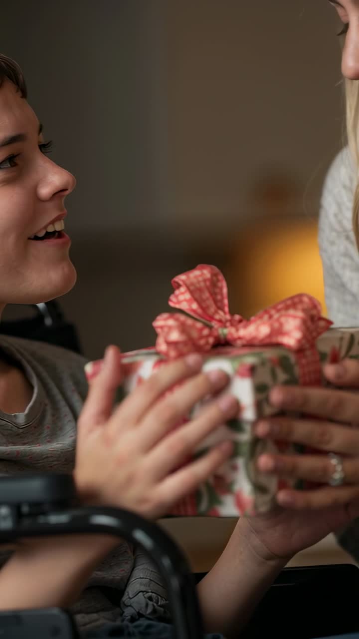 Video verticale: Bambino in sedia a rotelle che prende una scatola mentre la mamma con un maglione offre un regalo a casa, condividendo calore.