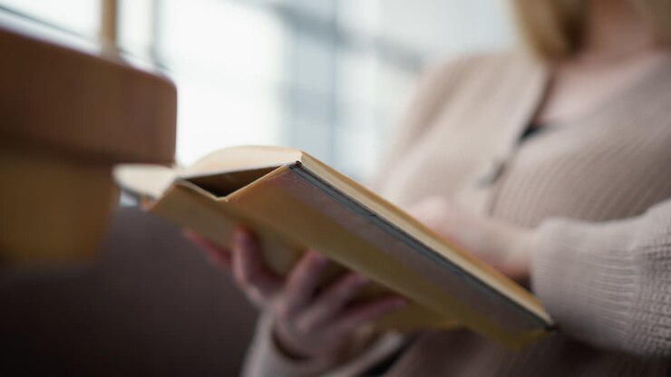 Primo piano di una giovane donna che tiene un libro mentre legge attentamente in un ambiente accogliente e rilassato; le sue dita sfogliano delicatamente le pagine mentre si immerge nella lettura.