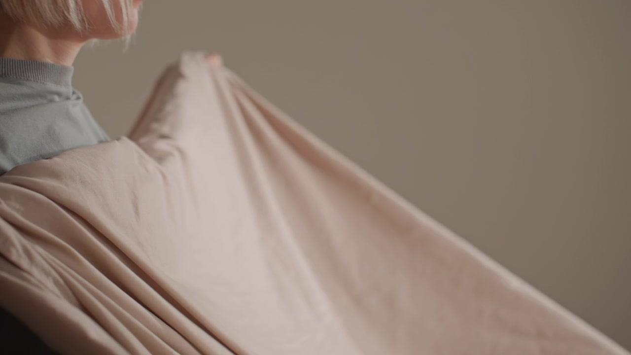White woman folding pale sheet, hands smoothing linen on bed, casual shirt and shorts, neutral studio bedroom, deliberate gestures, domestic chore rendered as tender ritual, quiet cozy atmosphere