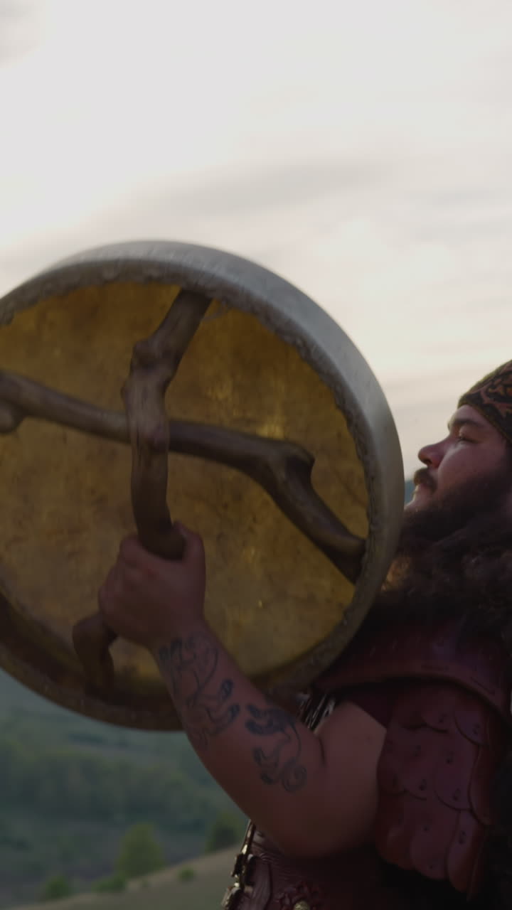 Modern shaman with fur cover plays drum with root handles in mountain valley. Sacred ritual of communication with spirits. Meditative sound