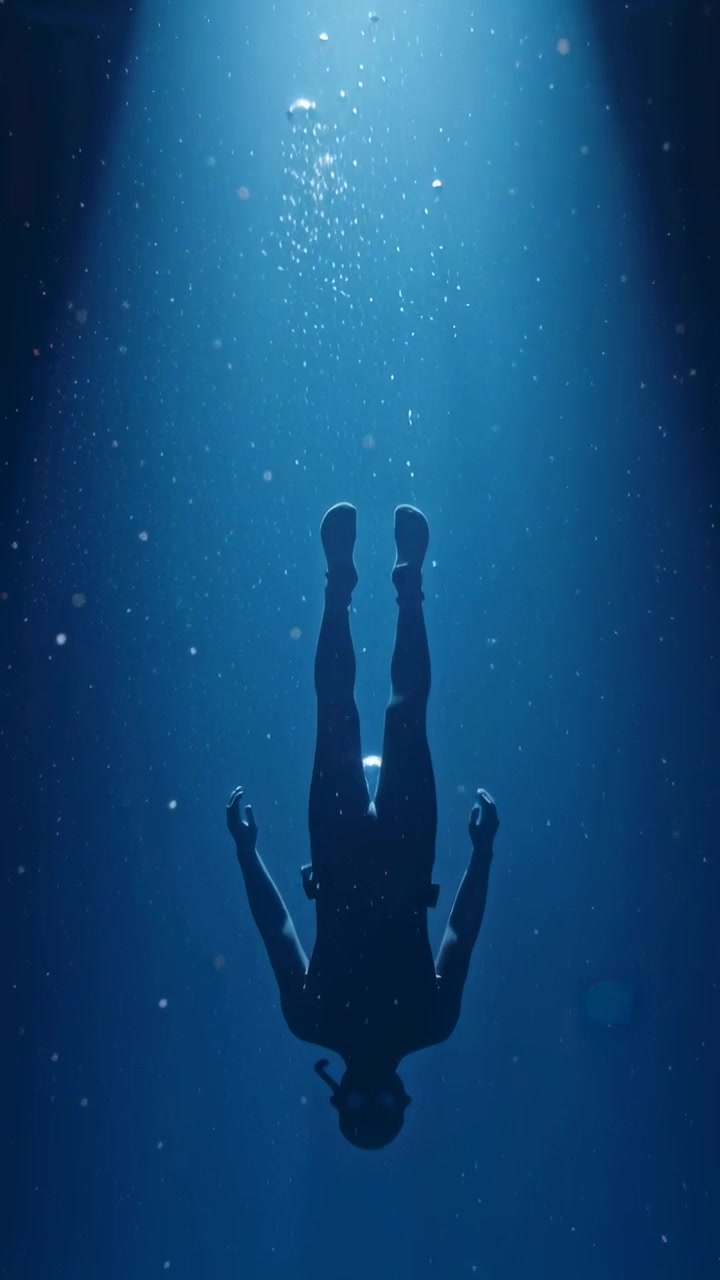 Video verticale: Subacqueo che scende in muta, con bombola e pinne, espirando bolle attraverso la luce nel mare profondo.
