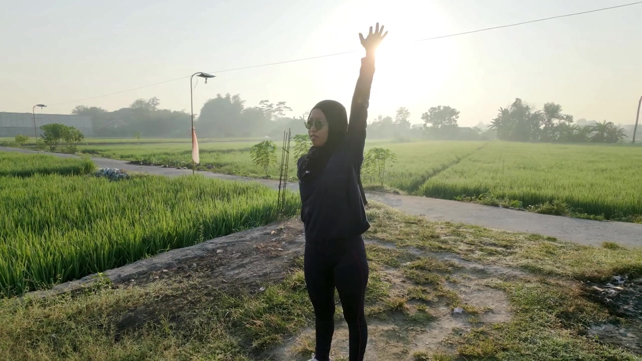 Pengambilan gambar orbit dinamis di sekitar seorang wanita Muslim berhijab dan berkacamata hitam yang sedang melakukan peregangan lengan untuk pemanasan. Sinar matahari pagi yang indah di sawah hijau yang menawan. Konsep kebugaran luar ruangan.