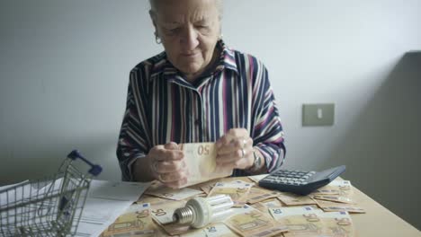 senior woman counting 50 euro note on desk with shopping cart and calculator light bulb paying bill utility