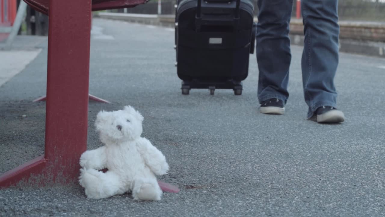 teddy abandonado en el andén del tren mientras un viajero pasa con una maleta en plano medio.