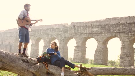 happy young couple backpackers tourists on a log trunk playing guitar singing in front of ancient roman aqueduct ruins in romantic parco degli acquedotti park in rome at sunrise slow motion