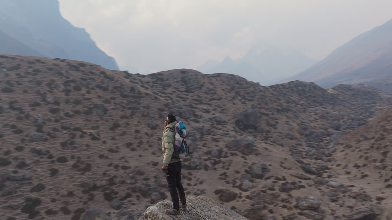 A tourist climbs a rock at Dingboche during Everest Base Camp trek. Drone hyperlapse captures wind and wide serene landscape of Himalayas experience of tourist in Nepal