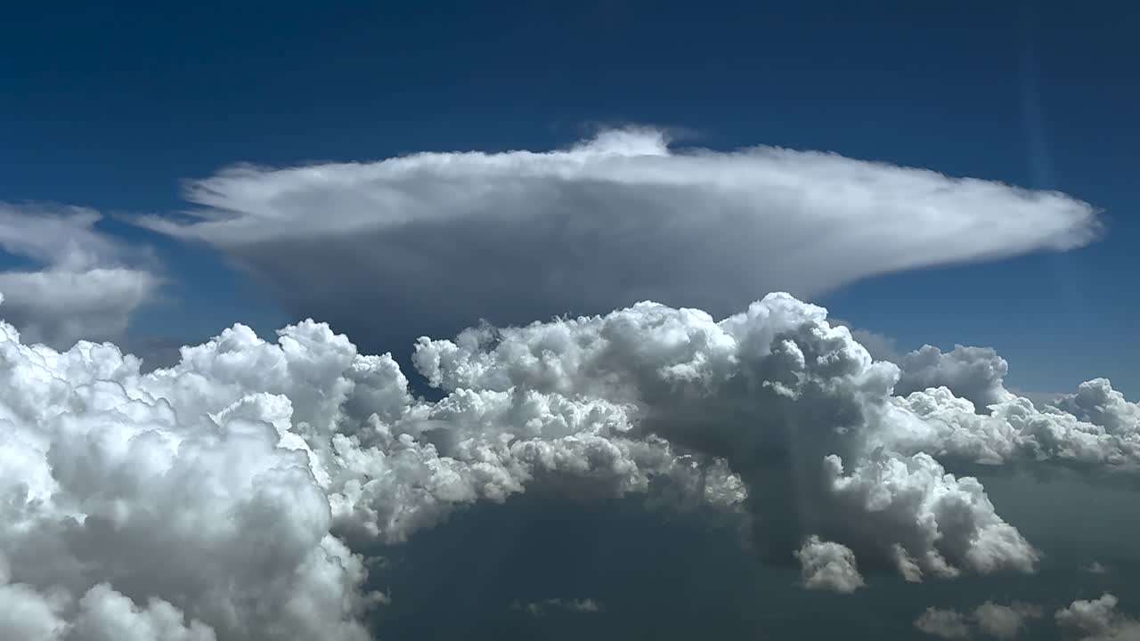 A stormy cloudscape from the sky, as seen by the pilot from a jet cockpit, with flying over cottony clouds, with a massive threatening cumulonimbus cloud at the back. Ultra-realistic 4k