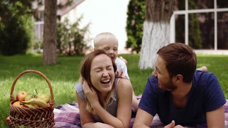 young, beautiful couple lies on a belly on a plaid in the park. have a rest on the nature. sunny day. a small son climbs on his mothers back, frolics, has fun. everyone is smiling. family idyll