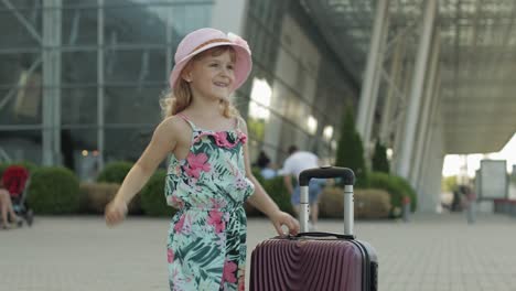 child girl tourist with suitcase bag near airport. kid with luggage dances, rejoices, show thumbs up