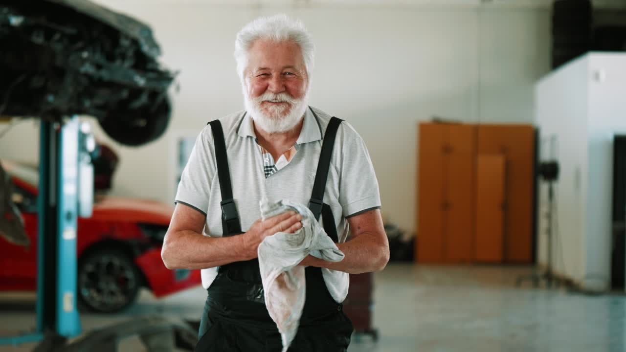 Confident old male mechanic cleans hands and looking at camera in  garage. Portrait