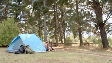 two women relax beside a blue tent in a wooded campsite with copy space