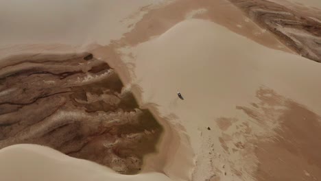 Aerial:-A-truck-with-kitesurfers-traveling-through-the-dunes-of-Lencois-Maranhenses-in-Brazil,-during-dry-season