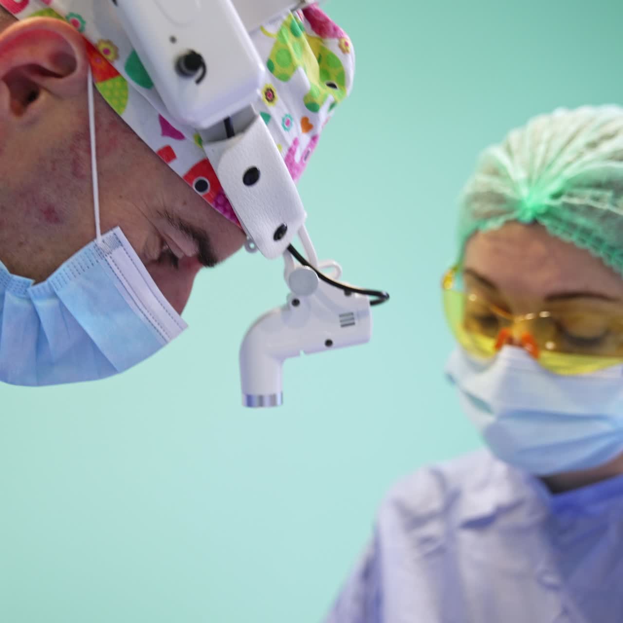 Side view of a surgeon in cap, mask and headlight who bent over the patient. Nurse in yellow protective goggles assisting the doctor
