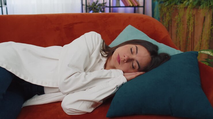 Tired young woman lying down in bed taking a rest at home, napping, falling asleep on orange couch