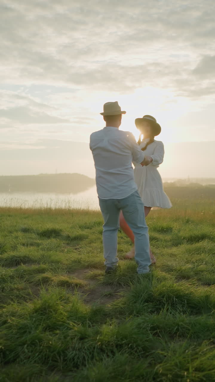 해가 지는 동안 잔디로 인 언덕 에, 평온한 호수 에서 사랑으로 회전하는 연인들은 색 셔츠, 모자, 청바지를 입은 남자가 색 드레스를 입은 아내를 포옹합니다.
