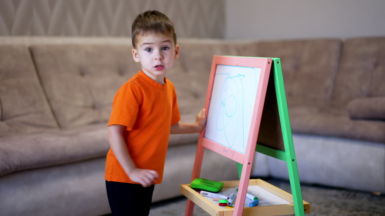 Adorable baby boy draws on the whiteboard with a blue marker. Toddler boy puts a marker open and walks away.