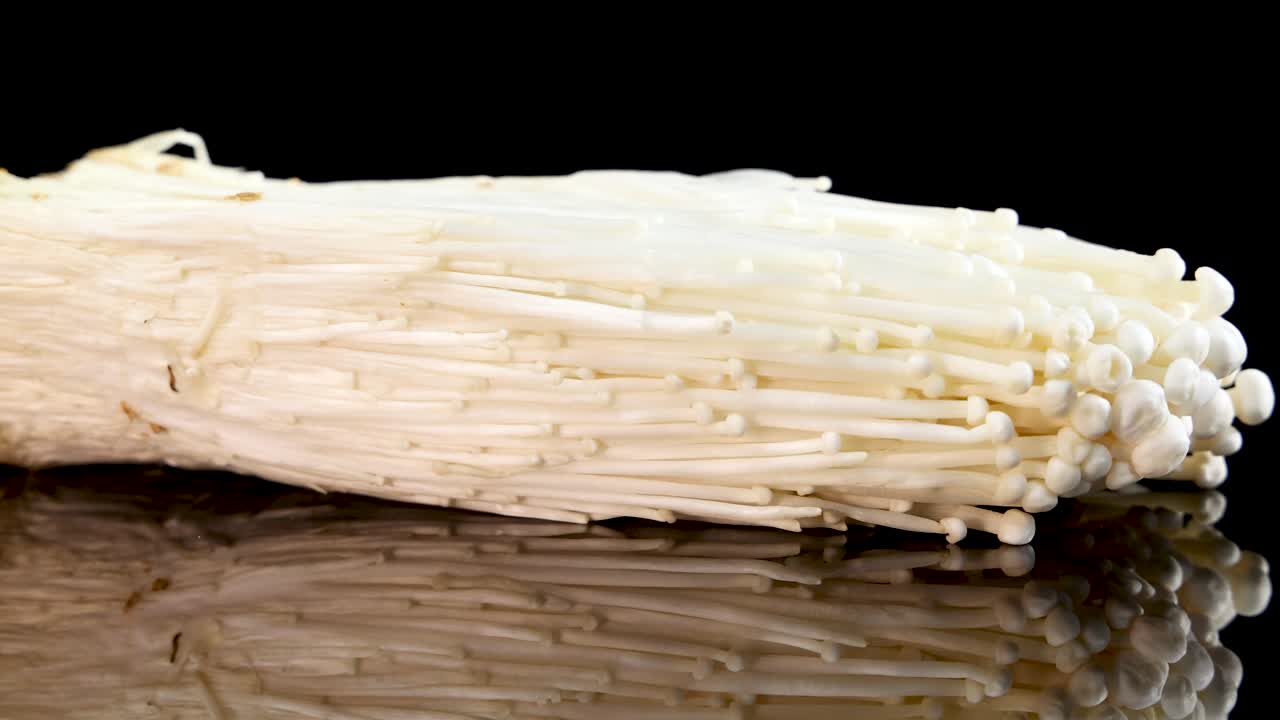 A bundle of enoki mushrooms slowly rotates on a glossy black surface under bright studio lighting, highlighting texture and delicate structure with clear reflections