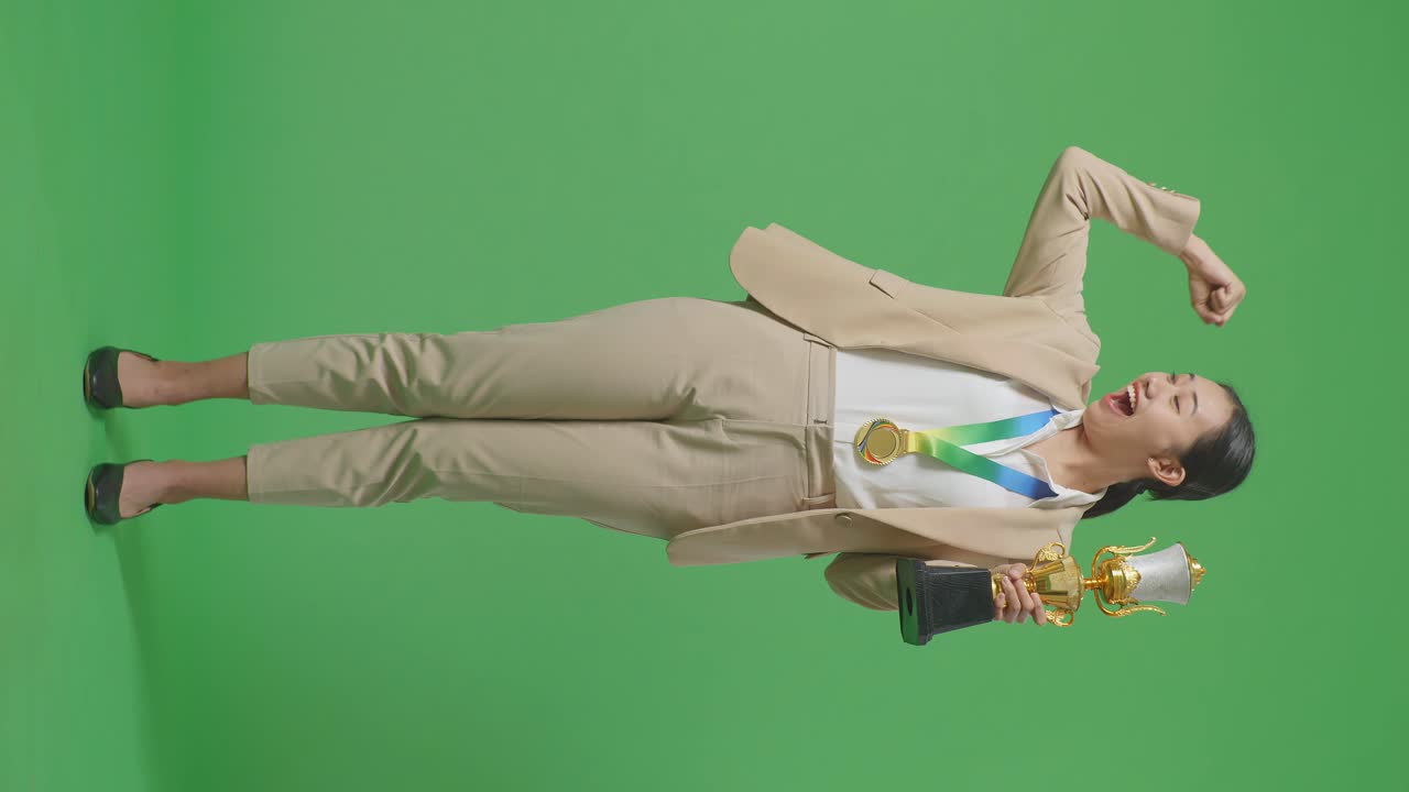 골드 메달과 트로피를 입은 아시안 사업가 여성의 몸이 가득 차 있으며 스튜디오의 녹색 화면 배경에서 카메라에 미소 짓고 있습니다.