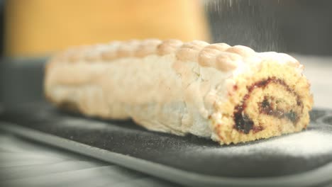 close up of a swiss roll with flambeed egg white cream while powdered sugar is put on top of the roll laying on a plate