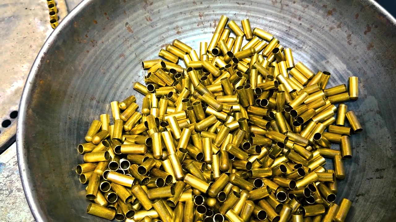 Golden brass bullet casings being sorted and agitated in a large metal bowl on an industrial production line at an ammunition factory, showcasing the manufacturing process of military ordnance