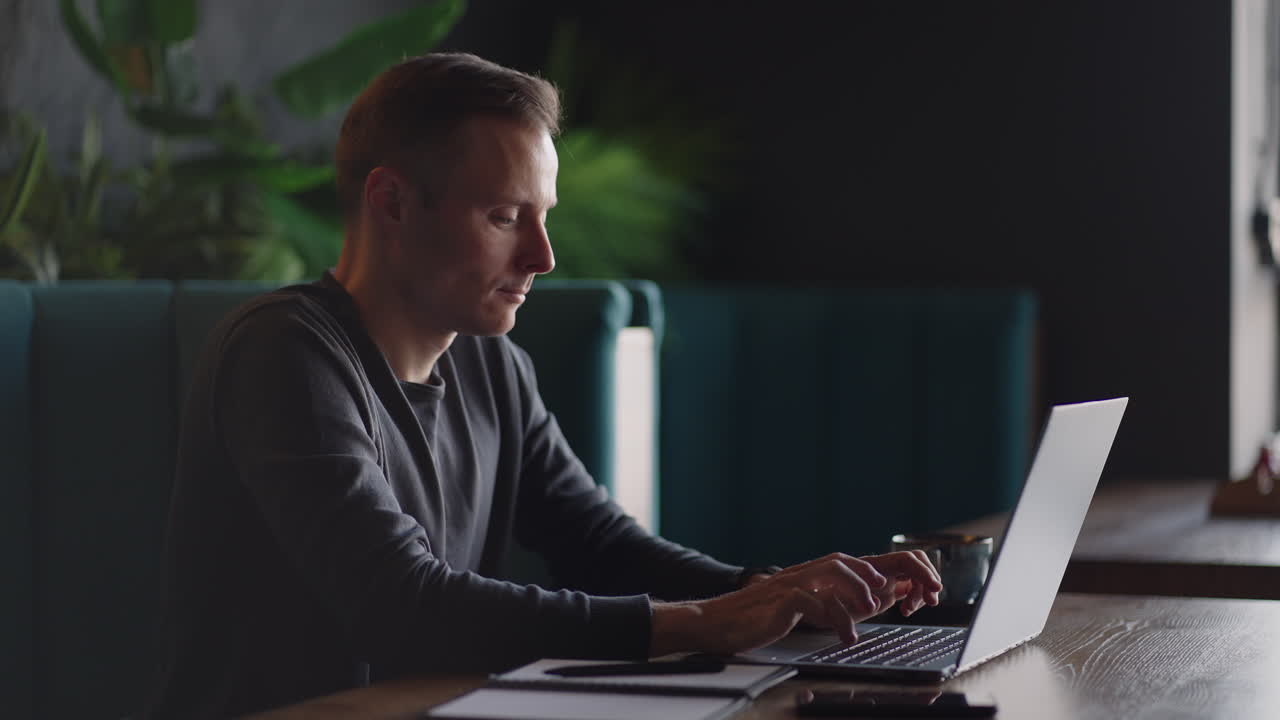 A young man in a shirt is sitting at a table with a laptop and typing on the keyboard. A student can study remotely. A businessman conducts his business remotely