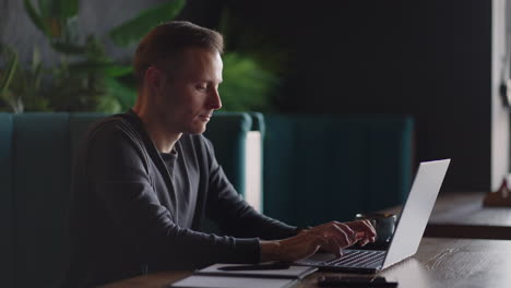 a young man in a shirt is sitting at a table with a laptop and typing on the keyboard. a student can study remotely. a businessman conducts his business remotely