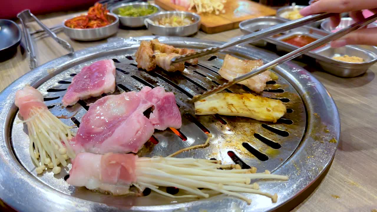 Hands grill meat and vegetables on a tabletop BBQ in a vibrant Bangkok setting. Warm lighting enhances the lively dining atmosphere
