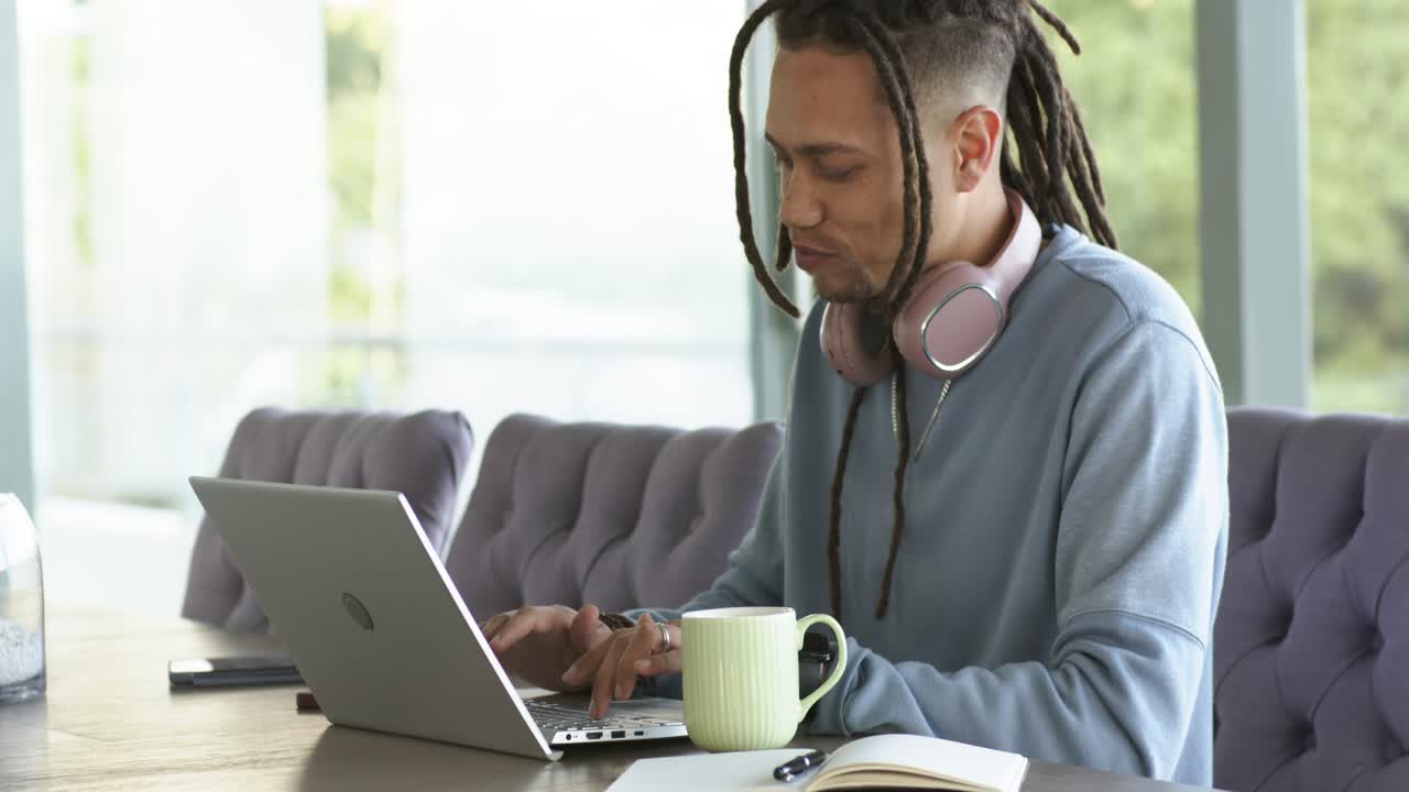 Afroamerikansk mand sidder derhjemme, iført lyserøde hovedtelefoner, og taster på en sølvfarvet bærbar computer for at arbejde.