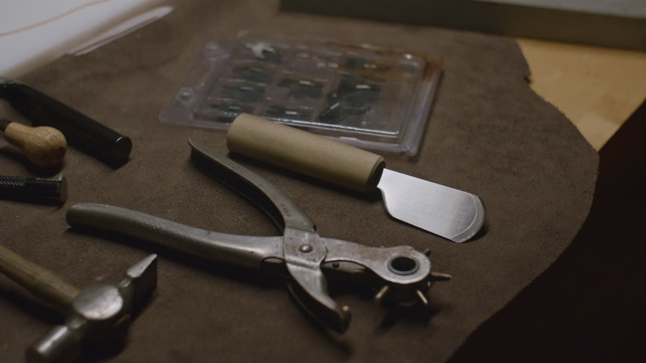 A collection of leatherworking tools including a hole punch, skiving knife, hammer, and chisels laid out on a brown suede work mat.