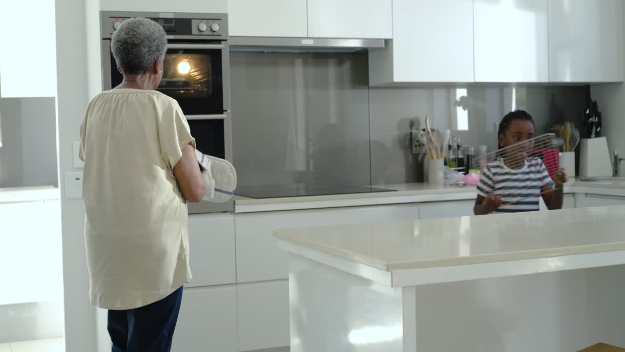 Anziana donna africana apre il forno con i guanti da cucina, mentre una giovane applaude e osserva il vassoio in cucina.