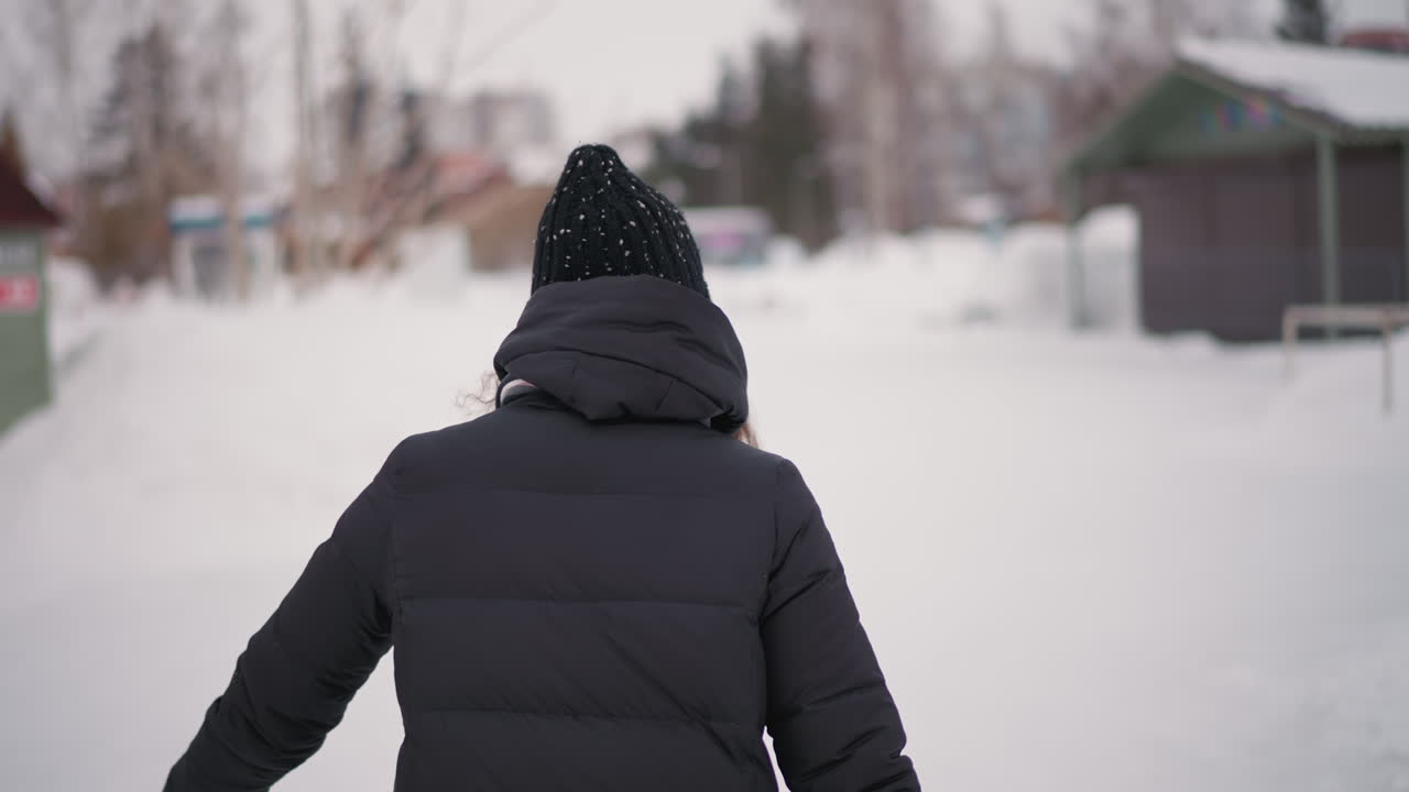 Athletic woman wearing black winter jacket, scarf, and knitted hat walks outdoors in snowy environment, curly hair visible from under hat, back view showing strength, resilience in cold weather scene