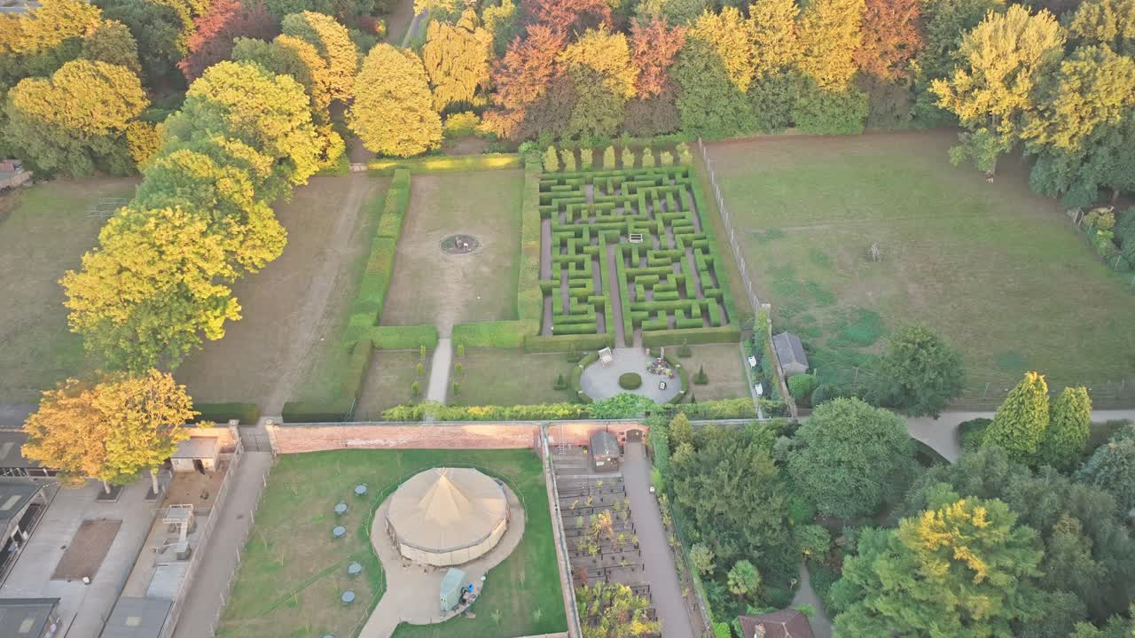 From a dolly-in drone motion, Wentworth Woodhouse’s hedge maze and circular pavilion emerge from tree-lined fields, with symmetrical paths and historic stone buildings framed by autumn shadows
