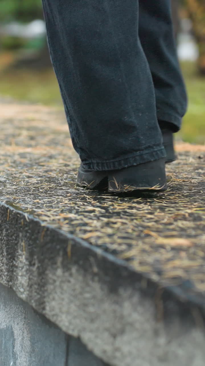 Back view of person wearing black trousers and boots carefully walking along wide wet stone path during rainy autumn day, with fallen pine needles scattered on ground and blurred park backdrop