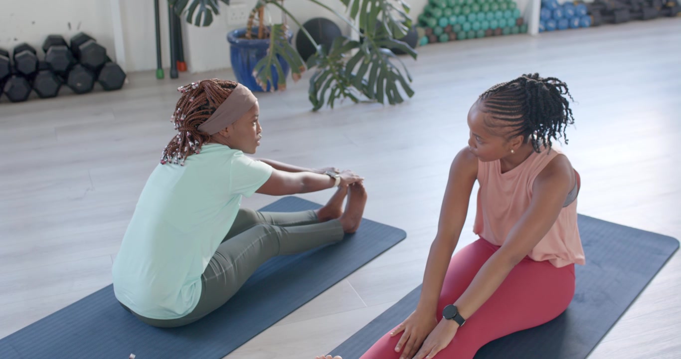 Afroamerikanische Frauen im Fitnessstudio auf Matten, linke Frau streckt sich zu den Zehen, ermutigt Partnerin zum Nachahmen für mehr Beweglichkeit