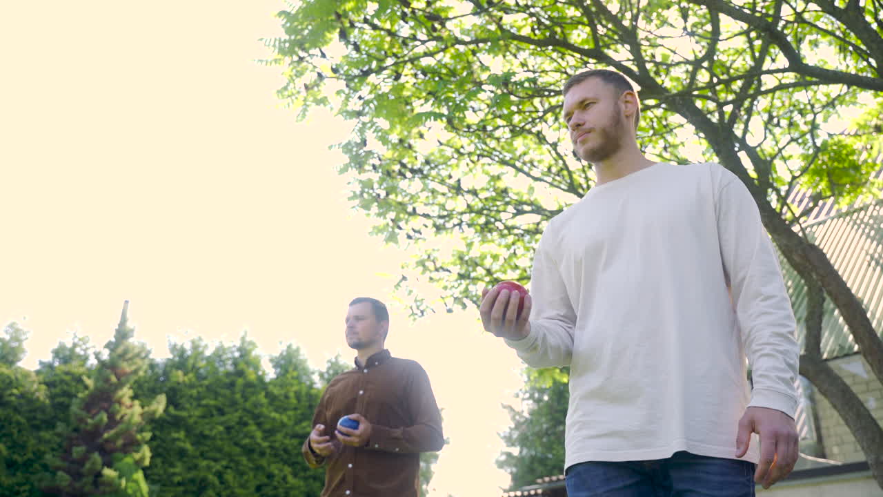 彼の男性の友人が彼の番を待っている間、晴れた日に公園で赤いペタンクボールを投げる白人の若い男の底面図