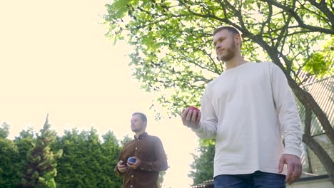 Bottom-view-of-caucasian-young-man-throwing-a-red-petanque-ball-in-the-park-on-a-sunny-day-while-his-male-friend-waits-his-turn