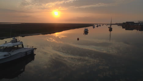 low drone shot over still water with small boats and sailing boats on creek at high tide with salt marsh and stunning sunrise in north norfolk uk east coast